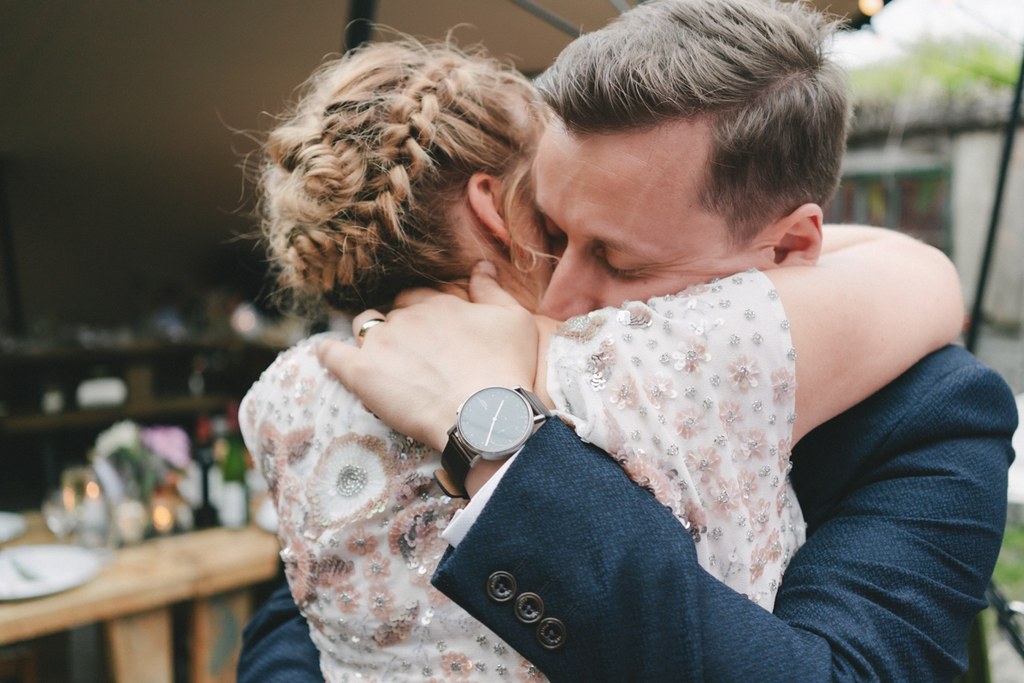 Groom in blue suit embraces the bride on the terrace — decorative flowers in the background