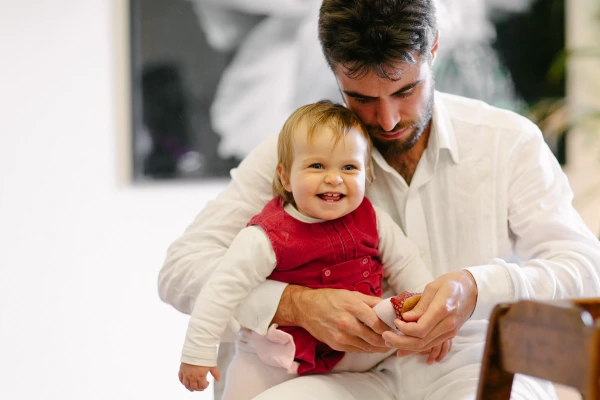 A bearded man in a white shirt holds up a grinning baby in a red dress at a wedding reception, feeding her a strawberry. Candid and joyful documentary wedding photograph.