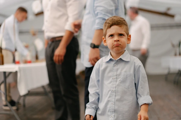 Young boy in light blue shirt at wedding reception with guests and white balloon decorations inside tent.