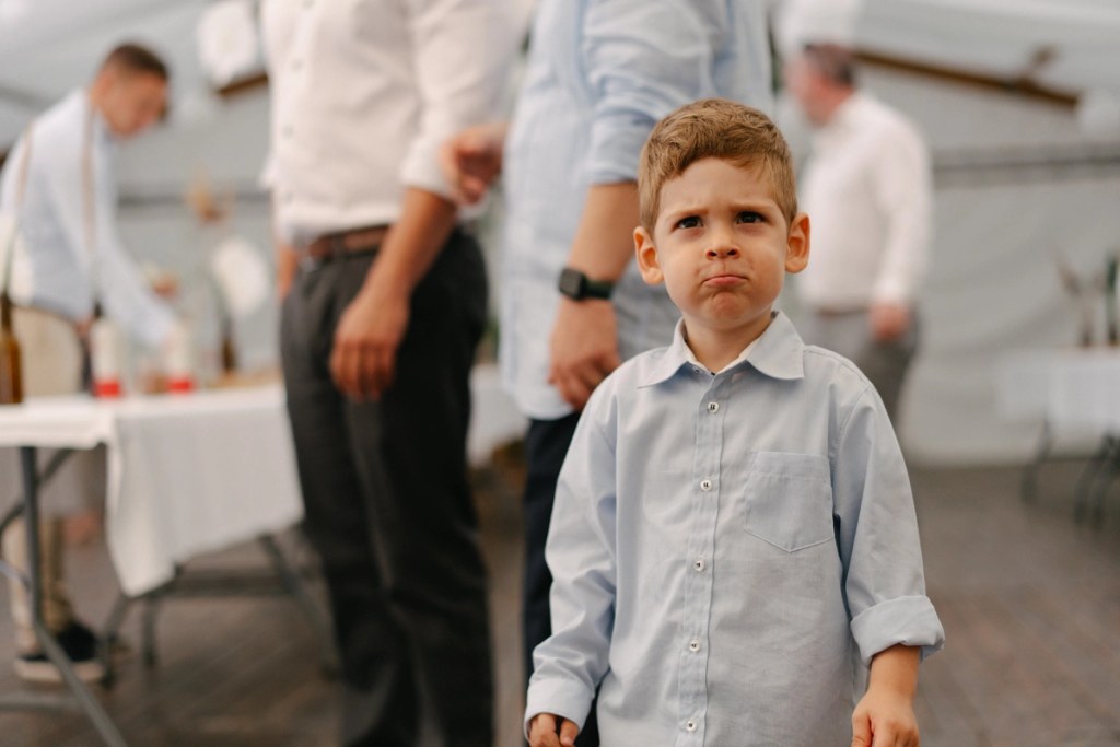 Young boy in light blue shirt at wedding reception with guests and white balloon decorations inside tent.