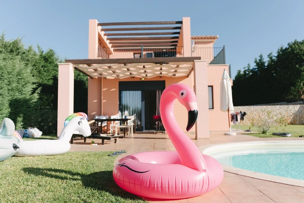 Inflatable flamingo float at the pool edge, terracotta house and pergola behind, clear blue sky