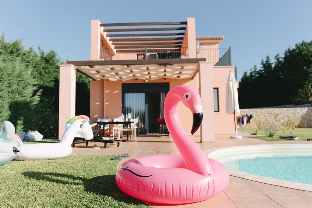 Inflatable flamingo float at the pool edge, terracotta house and pergola behind, clear blue sky
