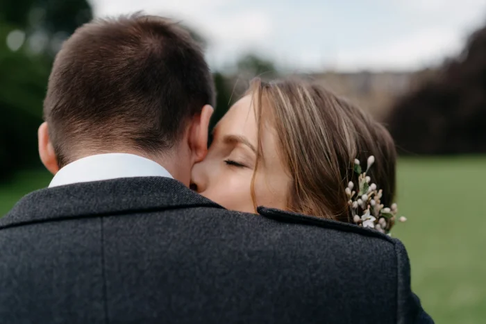 Bride with floral hair accessory embracing groom in dark suit outdoors, eyes closed, tender wedding moment on green lawn.