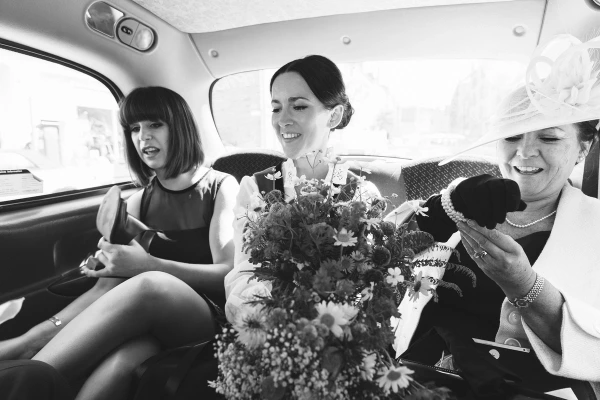 Black and white shot of the bride holding her wildflower bouquet in the back of a taxi between a bridesmaid and her elegantly hatted mother. Candid pre-ceremony moment.