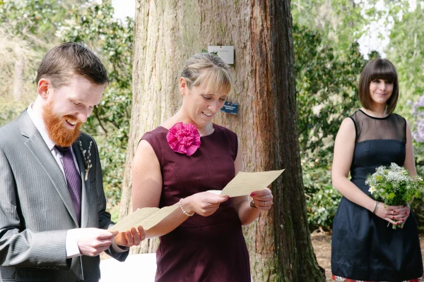 Candid documentary shot of a guest reading from a sheet of paper during the ceremony while a bridesmaid with a wildflower posy looks on. Outdoor reading at the Royal Botanic Garden Edinburgh.
