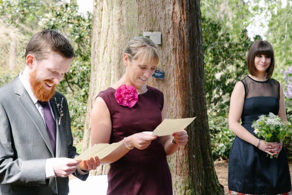 Candid documentary shot of a guest reading from a sheet of paper during the ceremony while a bridesmaid with a wildflower posy looks on. Outdoor reading at the Royal Botanic Garden Edinburgh.