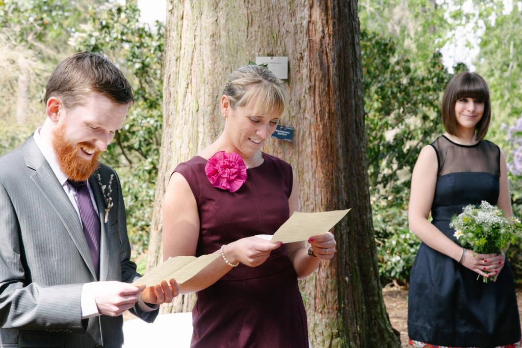 Candid documentary shot of a guest reading from a sheet of paper during the ceremony while a bridesmaid with a wildflower posy looks on. Outdoor reading at the Royal Botanic Garden Edinburgh.