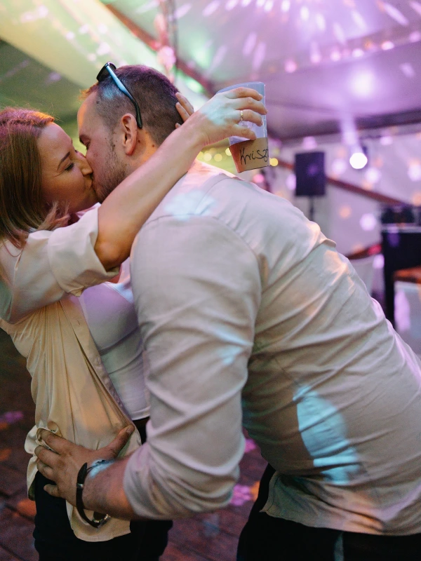 Couple kissing at wedding party — colorful ambient purple lighting, champagne glasses, intimate celebration moment.