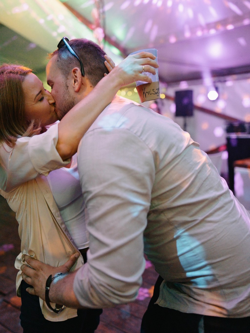 Couple kissing at wedding party — colorful ambient purple lighting, champagne glasses, intimate celebration moment.