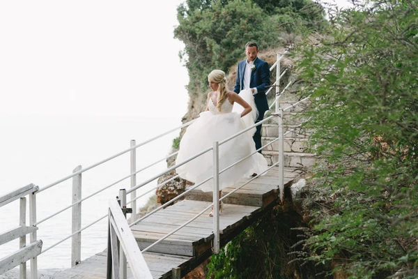 Bride in white lace and groom in dark blue walk on a wooden bridge by rocky seaside