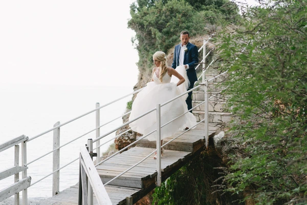 Bride in white lace and groom in dark blue walk on a wooden bridge by rocky seaside