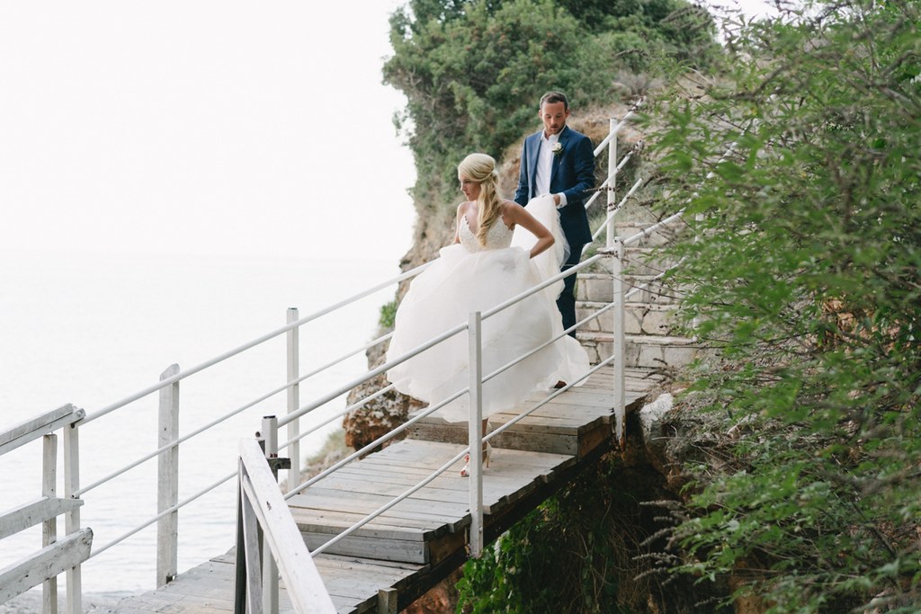 Bride in white lace and groom in dark blue walk on a wooden bridge by rocky seaside