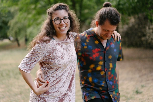 Woman in glasses smiling with arm around man in colorful patterned shirt at outdoor wedding venue.
