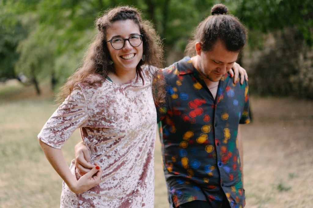 Woman in glasses smiling with arm around man in colorful patterned shirt at outdoor wedding venue.