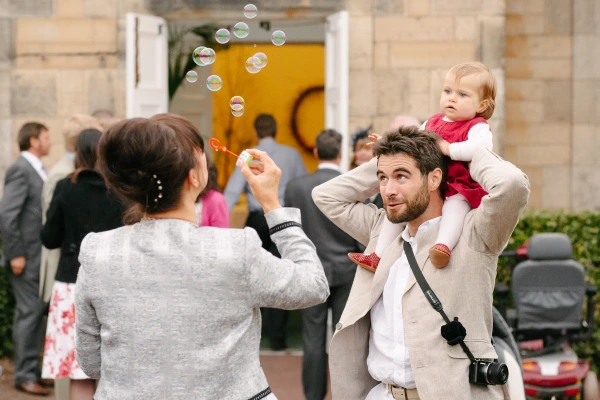 A father carries a baby in a red dress on his shoulders while blowing soap bubbles, captured in a lively candid moment outside the wedding venue. Documentary wedding photography.