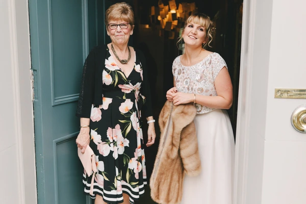 Two women in front of a turquoise door — older lady in black floral dress, younger in white lace. Elegant wedding moment.