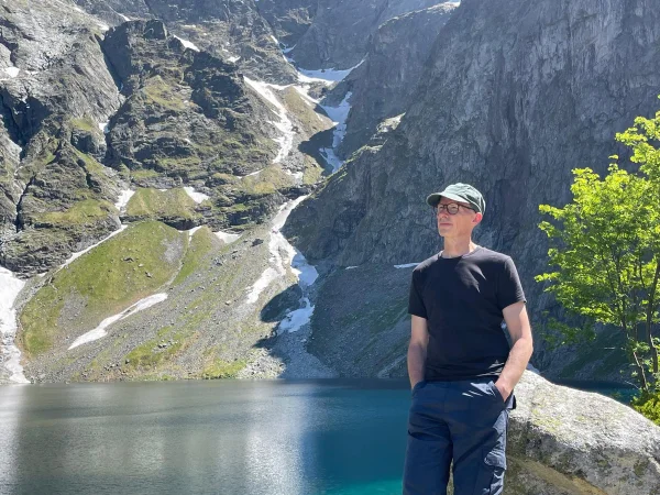 Istvan Magyar in black tee and green cap stands before a turquoise alpine lake surrounded by snow-streaked rocky cliffs.