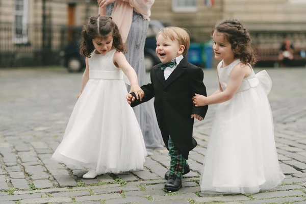 Three children playing on cobblestones at the wedding — white dresses, boy in tartan boots