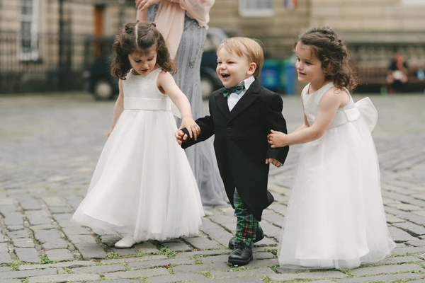 Three children playing on cobblestones at the wedding — white dresses, boy in tartan boots