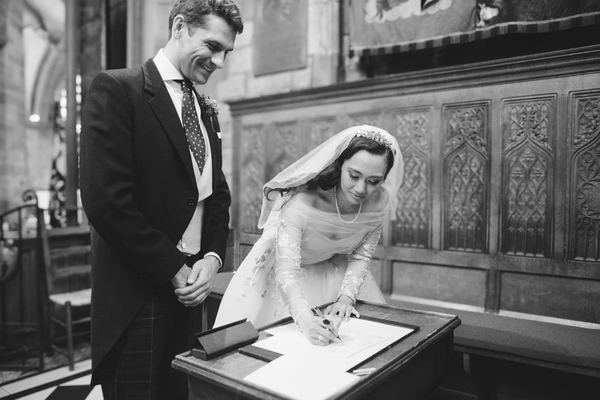Bride signs the marriage register, groom watches lovingly at the altar