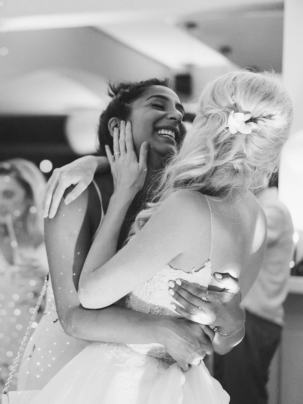 Bride with flower in hair hugs a female guest at the wedding — both smiling. Black-and-white.
