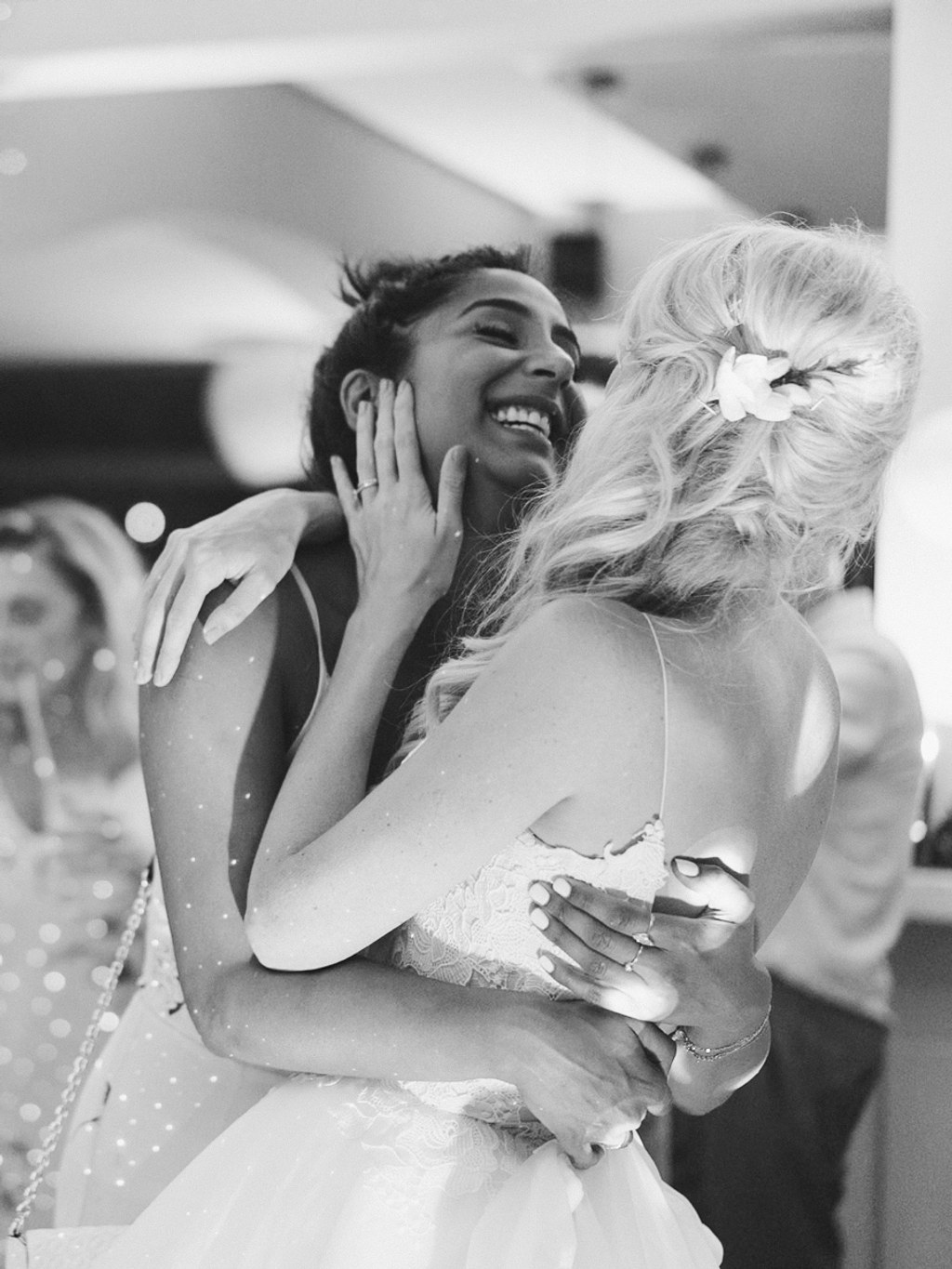 Bride with flower in hair hugs a female guest at the wedding — both smiling. Black-and-white.