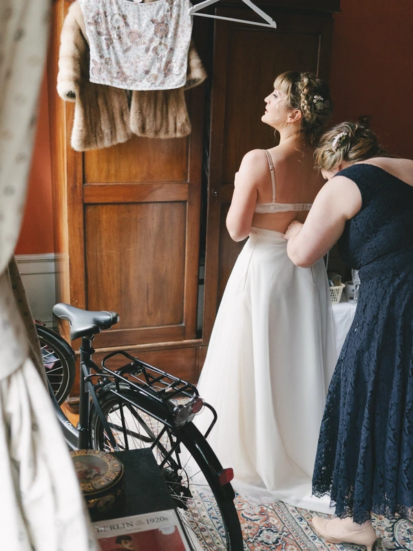 Bride in white dress with helper in a vintage room, white lace top hanging on the door above