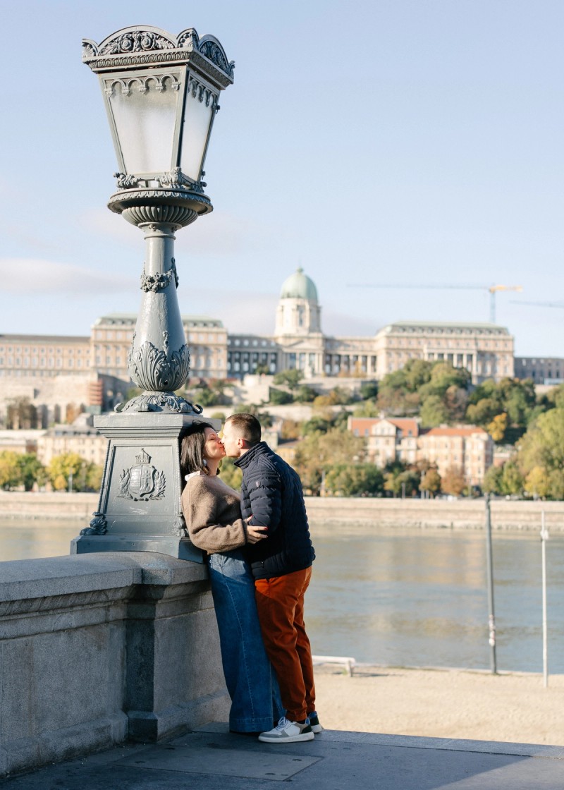 Jegyespár csókolózik Budapesten, a Lánchíd közelében egy kandelábernek támaszkodva a Duna-parton. A Budai Vár látható a háttérben.