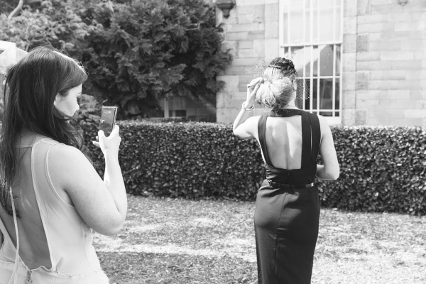 Black and white candid shot of a guest photographing her companion who adjusts her veil-style headpiece outside the venue. Playful documentary moment at the drinks reception.