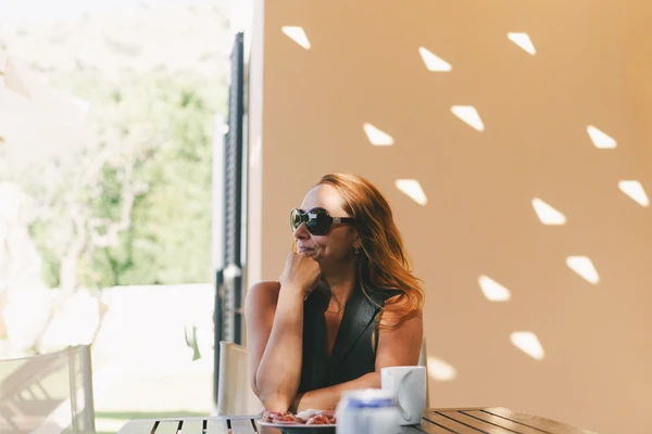 Woman in sunglasses and black shirt at a table, pensive look — latticed shadows on ochre wall