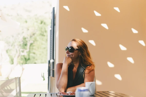 Woman in sunglasses and black shirt at a table, pensive look — latticed shadows on ochre wall