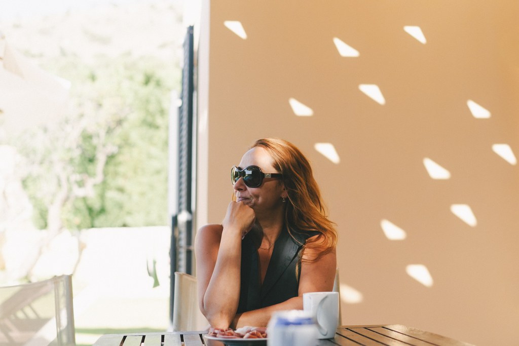 Woman in sunglasses and black shirt at a table, pensive look — latticed shadows on ochre wall
