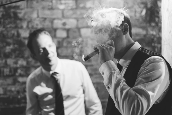 Man blowing cigar smoke, white-haired, watched by another man against a stone wall — black-and-white