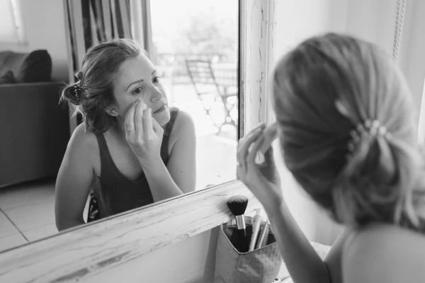 Bride looks thoughtfully at herself in the vanity mirror — brushes behind, natural window light