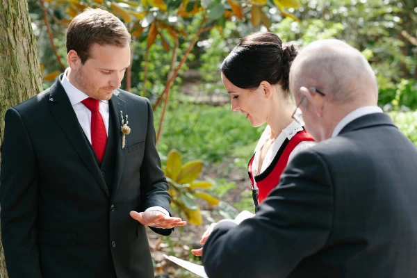Groom in a dark suit and red tie holding a green folded card as he reads his vows facing the bride, officiant and garden foliage in the background. Candid outdoor ceremony.
