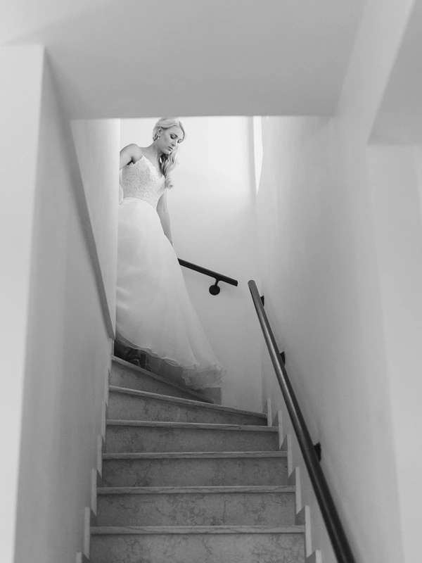 Bride descends the stairs in white lace, hand on banister — bright light behind, dramatic shadows