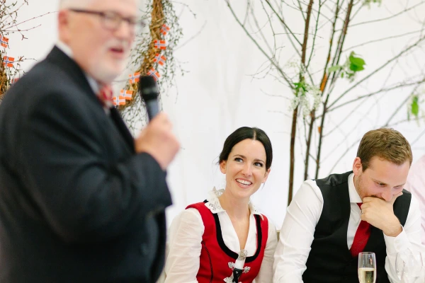 The bride in a Norwegian bunad smiles up at an older man giving a speech into a microphone at the wedding reception, with Scandinavian flags visible in the background. Candid documentary photograph.