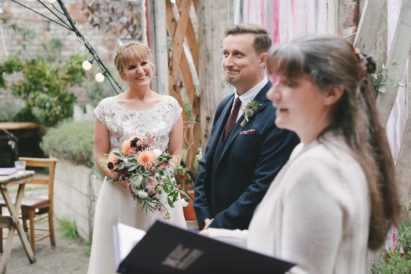 Bride in lace holds an orange bouquet, joyfully looking at her groom at the outdoor ceremony