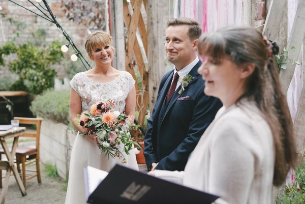 Bride in lace holds an orange bouquet, joyfully looking at her groom at the outdoor ceremony
