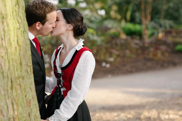 The groom in a dark suit and the bride in a Norwegian bunad share a kiss beside a tree in close-up, warmly lit by evening sun. Documentary wedding portrait at the Royal Botanic Garden Edinburgh.