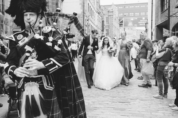 Scottish piper leads the bridal procession down a narrow street — black-and-white
