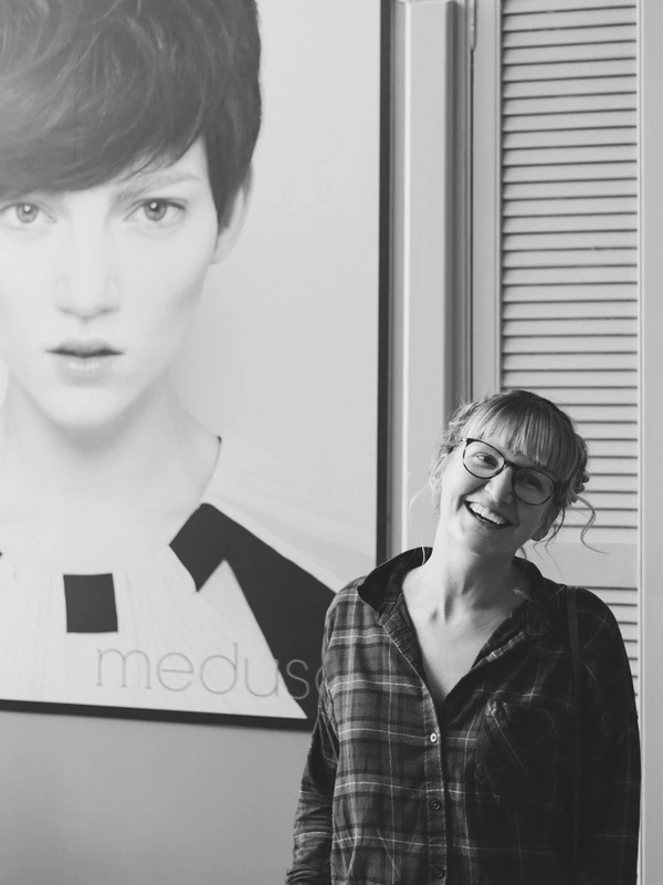 Young woman in glasses smiling at the camera, standing beside a black-and-white portrait
