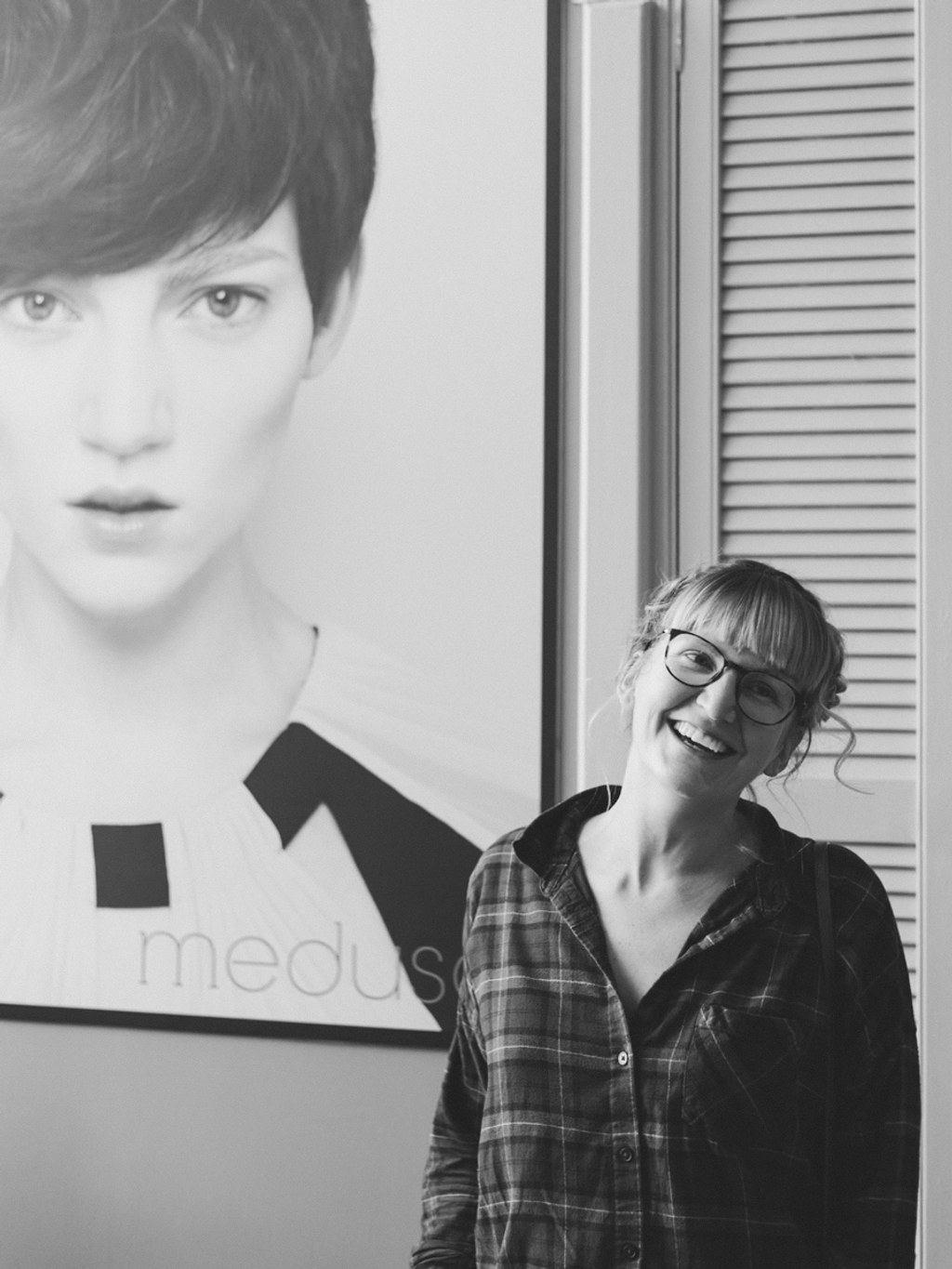 Young woman in glasses smiling at the camera, standing beside a black-and-white portrait