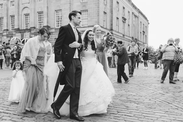 Bride and groom walk together before an imposing classical building, smiling, guests around them