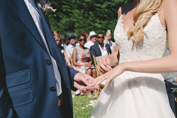 Bride and groom exchange rings at the ceremony before a green hedge, guests watching in sunny garden