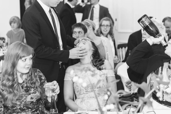 Black and white image of a wedding guest throwing his head back in laughter while a magician or entertainer performs at the reception table, as another guest pours wine. Candid documentary wedding moment.