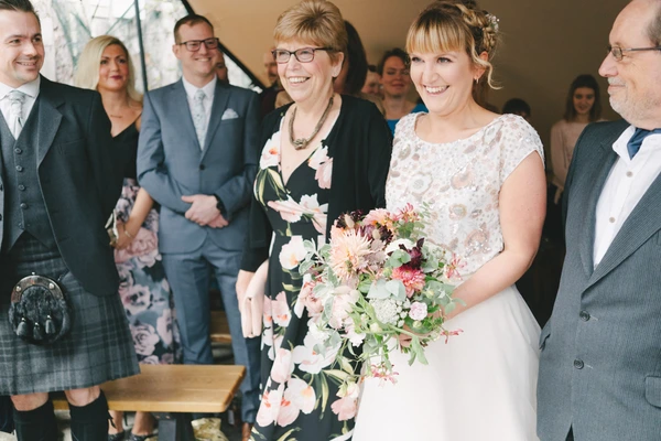 Smiling bride in white lace dress with pink and burgundy bouquet surrounded by suited guests