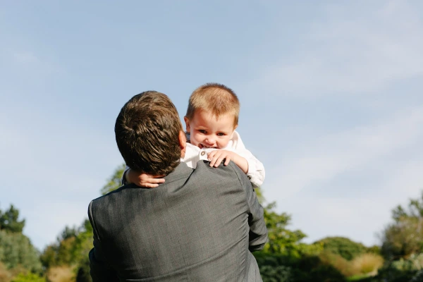 A man in a grey suit holds a smiling baby boy on his shoulder against a bright blue sky during the wedding reception at the Royal Botanic Garden Edinburgh. Candid documentary moment.