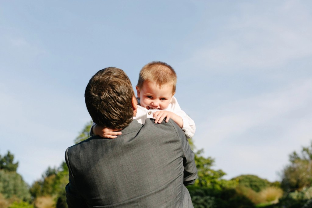 A man in a grey suit holds a smiling baby boy on his shoulder against a bright blue sky during the wedding reception at the Royal Botanic Garden Edinburgh. Candid documentary moment.