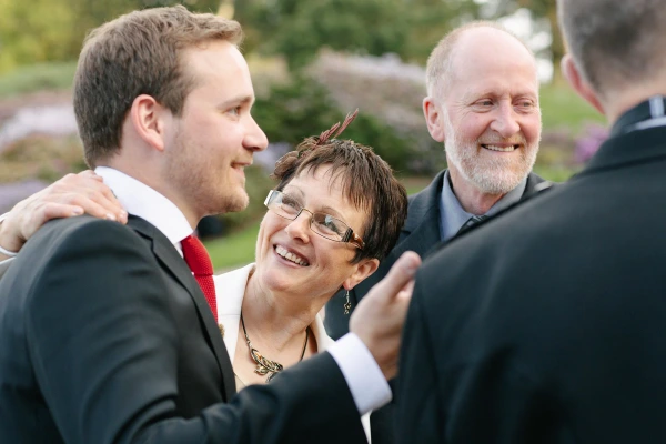 The groom shares a warm moment with his mother and father during the drinks reception in the garden, all three laughing together. Candid documentary wedding photography at the Royal Botanic Garden Edinburgh.